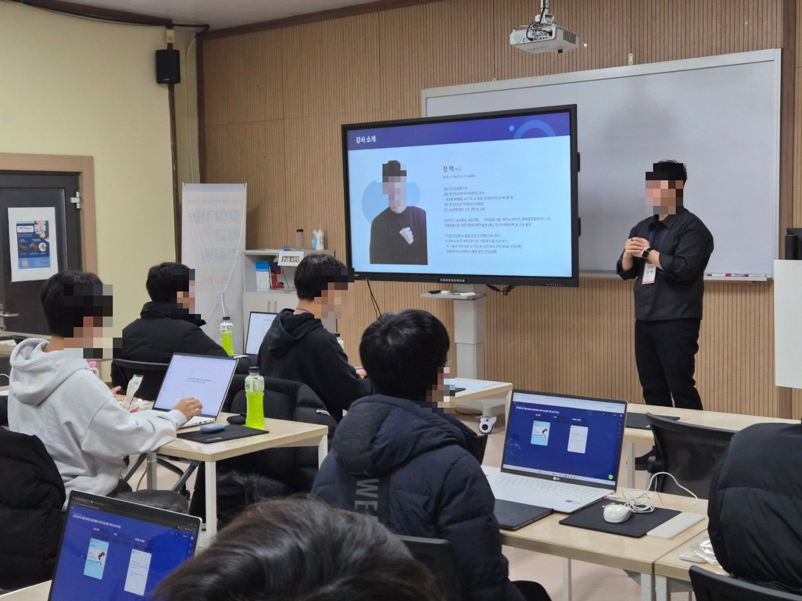 강의실에서 발표자가 강사 소개 화면 앞에서 수업하는 장면